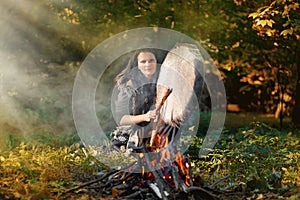 Shaman woman drumming next to the fire in the forest in the setting sun