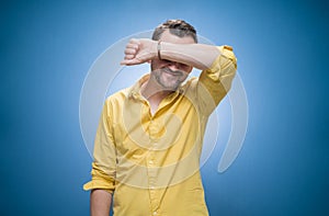 Shame. Young man covering face with hands over blue background, dresses in yellow shirt. Ashamed guy