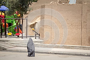 Shape of a woman walking in the streets of Yazd, Iran, passing near a children playground, wearing a niqab