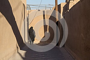 Shape of a woman walking in the streets of Yazd, Iran, passing near a traditional clay walls of the old city, wearing a niqab