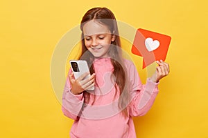 Sharing popular content on social media. Smiling little girl wearing pink sweatshirt holding blogger heart like icon and using