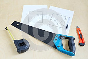 Sharp hacksaw, tape measure and knife on wooden surface, building tools next to blueprint