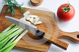 Sharp knife, vintage solid oak wooden cutting board, chopped green onion and fresh tomato on a light background. The concept of
