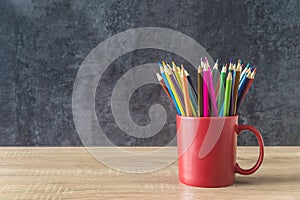 Sharpend colorfull pencils in red mug on a wooden desk against gray background