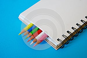 Sharpened colored pencils lie together with a blank notepad on a blue background