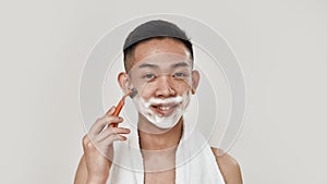 Shave easily. Portrait of shirtless young asian man with towel around his neck shaving his face, looking at camera