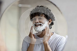 Shaving routine concept. Indian man applying shave foam on face making morning hygiene and looking at his reflection