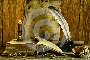 A sheet of old paper with space for text, an open book and a burning candle, an inkwell with a feather on a wooden background