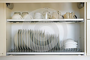 Shelf for tableware drying in modern kitchen. White glass and ceramic plates and cups on metal rack inside kitchen cupboard