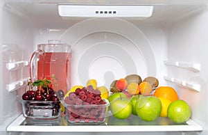 On the shelf of a white refrigerator, fresh fruit, berries and a glass jug with compote