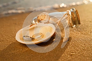Shell with gold wedding rings and invitation in glass bottle on sandy beach at sunset, closeup