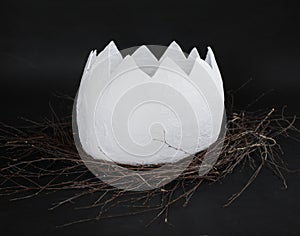 A shell of a huge papier mache egg in a nest of branches on a black background