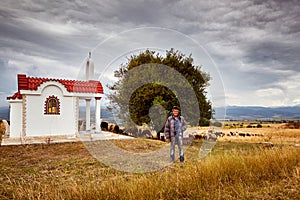 A shepherd in front of a little chapel and a herd of sheep