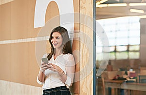 Shes always connected. a smiling young woman using her cellphone while standing in an office.