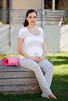 Shes still doing yoga every day. a young pregnant woman sitting with her yoga mat outside.