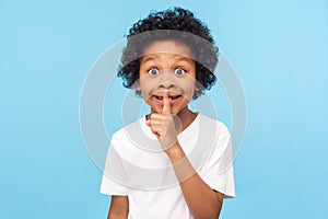 Shh, be quiet! Portrait of funny cute little boy with curly hair in T-shirt making silence gesture with finger on his lips