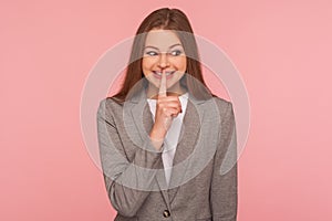 Shh, don`t tell anyone! Portrait of happy young woman in business suit smiling and holding finger on lips