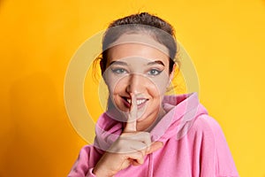 Shh. Keeping secrets. Portrait of young beautiful girl posing in pink hoodie over yellow studio background. Concept of
