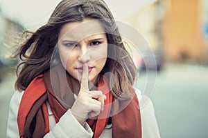 Shh Woman wide eyed asking for silence or secrecy with finger on lips hush hand gesture cityscape outdoor background Pretty girl