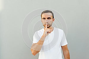 Shhh gesture. Young man dressed in white casual t-shirt pressing finger to lips as if asking to keep silence isolated on gray back