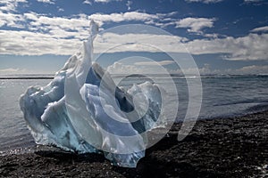 Shinny piece of ice over the black sand of Diamond beach, clouds on blue sky. Jokulsarlon - Iceland