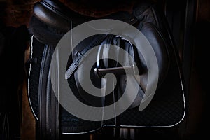 Shiny black leather horse saddle in the dark stable , with stirrup