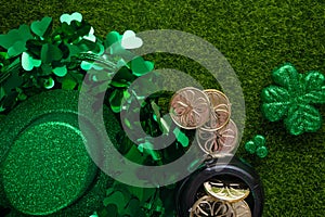 Shiny hat of leprechaun with clover and coins on green grass background. St. Patrick`s Day celebration. Flat lay. View from above