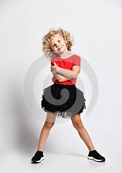 Shiny little kid girl in black skirt and red t-shirt stands with her legs wide hugging herself