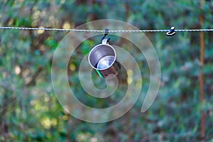 A shiny metal mug is hanging on a hook in the woods by a tree. A rope is stretched between the trees around the tent camp. Tourist