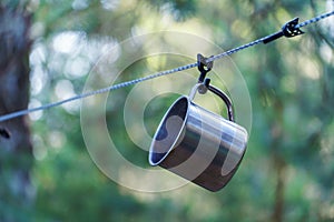 A shiny metal mug is hanging on a hook in the woods by a tree. A rope is stretched between the trees around the tent camp. Tourist