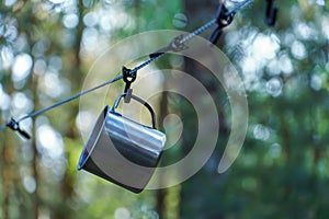 A shiny metal mug is hanging on a hook in the woods by a tree. A rope is stretched between the trees around the tent camp. Tourist