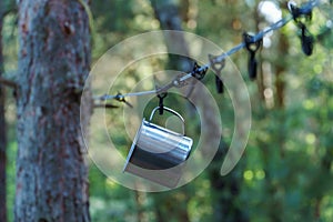 A shiny metal mug is hanging on a hook in the woods by a tree. A rope is stretched between the trees around the tent camp. Tourist