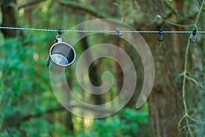 A shiny metal mug is hanging on a hook in the woods by a tree. A rope is stretched between the trees around the tent camp. Tourist