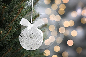 Shiny white ball with bow hanging on Christmas tree against festive lights, closeup. Space for text