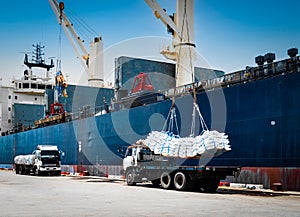 Ship crane lift-off slings of sugar bags cargo from truck and load into ship hold at seaport terminal.