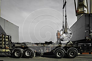 Ship crane put away steel coil on the trailer, worker stands on truck placing coil on the support skit.