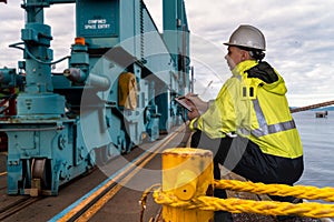 Ship supervisor engineer inspector stands at the dockside in a port. Wearing safety helmet and yellow vest. Cargo shipping