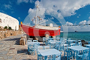 Ship and tavern furniture on quay in Mykonos, Greece. Red boat and blue tables on sea beach. Beach restaurant with sea