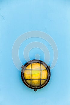 Ship yellow bulkhead light or deck lamp surrounded by a metal rusted frame fixed to a painted light blue pastel color photo