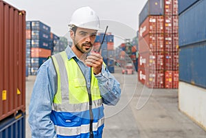 Shipment worker with safety vest and hardhat standing with walkie talkie in his hand. A large steel cargo containers stacked in