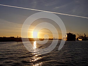 Shipping boat pulled by tugboats out of harbor as Shipping boat is unloaded by cranes in Oakland Harbor at sunset