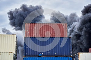Shipping containers stacked in storage with plumes of black toxic smoke from a fire against a blue sky.