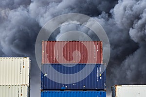 Shipping containers stacked in storage with plumes of black toxic smoke from a fire behind.