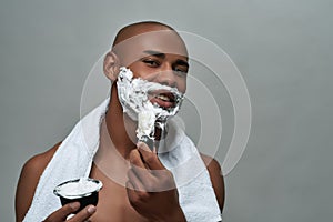 Shirtless attractive young african american man smiling at camera, using brush while applying shaving foam on his face