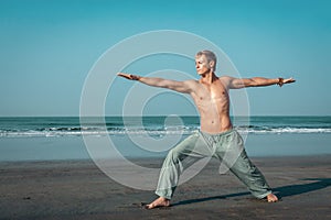 A shirtless, fit man stands in a wide warrior pose on the beach, with his arms outstretched and the ocean behind him