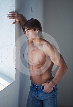 Shirtless handsome male with a perfect muscular body leaning on a wall in the studio, looking at a window.