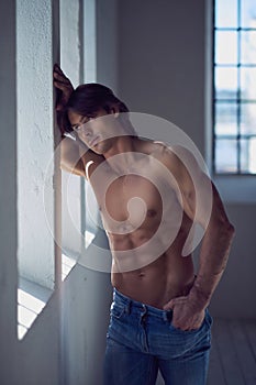 Shirtless handsome male with a perfect muscular body leaning on a wall in the studio, looking at a window.