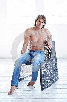 Shirtless handsome male with a perfect muscular body sitting on a chair in a studio.