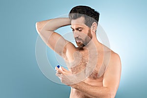 Shirtless handsome man spraying deodorant under arm for fragrance, hygiene and fresh scent, blue background