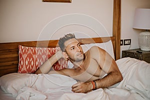 Shirtless male model lying alone on his bed in his bedroom, looking away with a seductive attitude.Carefree guy